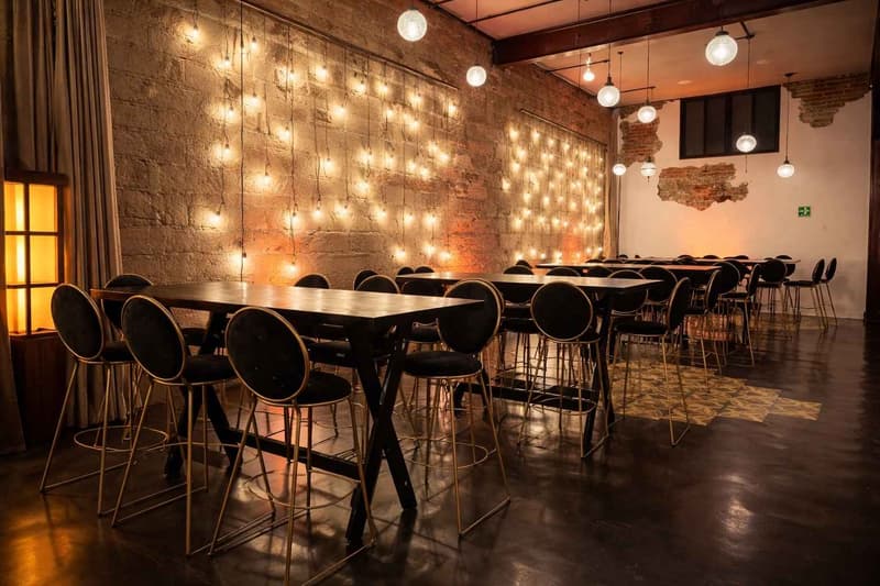 Montaje tipo bar interior con mesas altas y bancos de diseño, ambientado con iluminación cálida tipo guirnalda sobre muros de estilo industrial, ideal para cocteles y networking en eventos corporativos o sociales